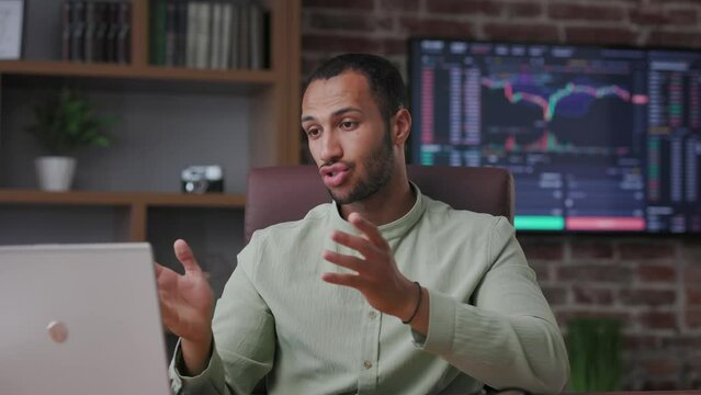 Smiling stock market broker talking to web camera sitting in office. African american trader has online video conference, video call, trading courses, recording vlog about cryptocurrency investment.