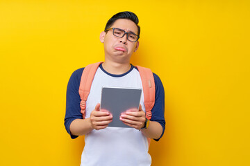 Displeased young Asian man student in casual clothes and glasses backpack holding digital tablet...