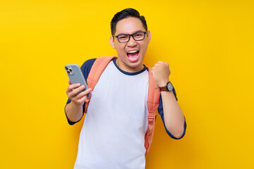 Excited young Asian man student in casual clothes backpack glasses holding mobile cell phone and saying yes, celebrating success isolated on yellow background. high school university college concept