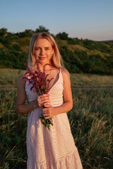 Smiling blonde young woman with wildflowers on nature background in summer at sunset. female is relaxing in the field with flowers. Soft golden color