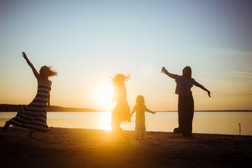 Fototapeta premium Silhouettes of group people dancing at sunset. Children and mothers jumping and having fun on the beach. Good mood. Active family pastime. Generations. Beautiful landscape. Enjoyment atmosphere