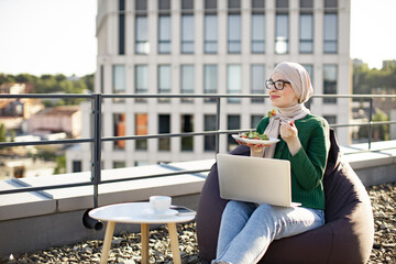 Calm muslim female wearing headscarf and glasses sitting in beanbag chair with vegan dish and...