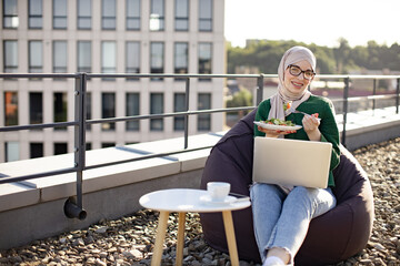 Young arabian woman in hijab holding phone while sitting near coffee table with laptop during lunch break on flat roof. Beautiful lady checking contact information before having bite of fresh salad.