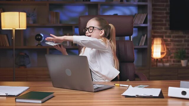 Energetic junior executive sitting in rolling chair and showering cash in trendy office space. Overjoyed blonde child with two ponytails celebrating company's huge financial income.