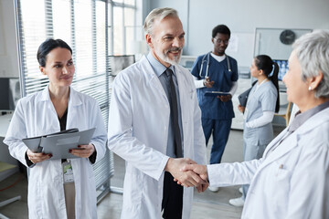 Smiling experienced hospital worker shaking hand of mature female colleague while congratulating...