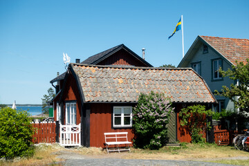 Fototapeta na wymiar Typical falu red house in Dweden with white corners on a picturesque rural historic fishing village island in Stockholm archipelago with Swedish flag against blue sky and the sea in the background