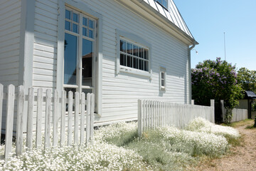 Idyllic white house with picket fence and small white flowers in front garden by entrance glass door with bright blue sky with peaceful rural small town feeling