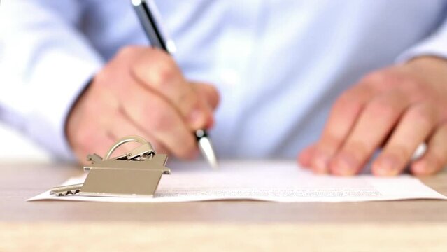 Close-up of hands of an unrecognizable caucasian man signing a home purchase contract with a pen. A house-shaped keyring is in focus.