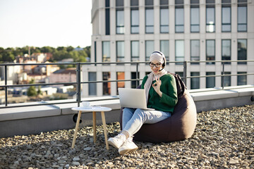 Relaxed muslim woman in headscarf conducting web conference via laptop and headset while staying on flat roof. Professional entrepreneur working outside of corporate offices via wireless devices.