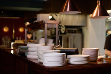 Several stacks of white plates standing on counter or workplace of chef surrounded by kitchen equipment and lamps hanging above