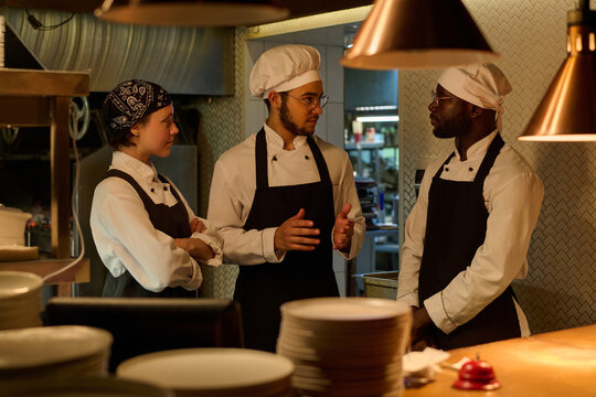 Three young intercultural cooks in uniform discussing working points in the kitchen while one of them explaining something to colleagues