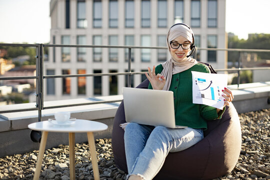 Focused muslim adult pointing at paper infographics during online meeting held outdoors via modern technologies. Calm businesswoman in hijab discussing financial data plan on panoramic terrace.