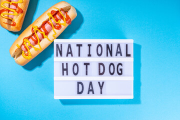

National Hot Dogs day background, hotdog summer party festival foods, Two tasty classic american hot dogs with sauces and lightbox sign with inscription National Hot Dog Day on blue background 
