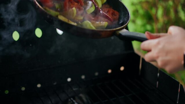 Hands shaking vegetables in frying pan over barbecue grill with smoke in slow motion. Unrecognizable Caucasian person cooking delicious picnic food outdoors