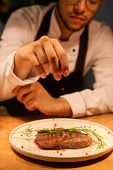 Focus on hand of young man in uniform sprinkling appetizing beef steak with spices to enrich taste of dish while standing by counter