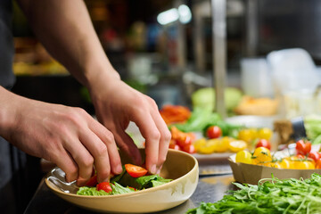 Hands of young male cook or chef putting fresh chopped ingredients of vegetable salad into plate while preparing dish for serving on table
