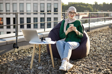 Fototapeta premium Full length view of charming woman in headscarf holding phone while sitting in beanbag near coffee table with gadgets. Delighted muslim person adoring city view in sunlight on panoramic terrace.