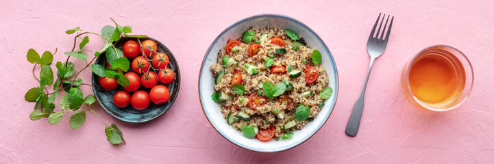 Quinoa tabbouleh salad in a bowl panorama, a healthy dinner with tomatoes and mint, with a drink, overhead flat lay shot on a pink background