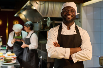 Happy African American male chef in uniform standing in front of camera in restaurant kitchen and...