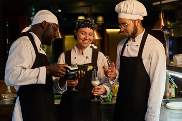 Young African American male chef pouring champagne into flutes held by his two colleagues during celebration of birthday of their restaurant