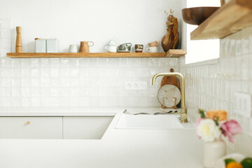 Modern white kitchen with brass faucet, granite countertop, wooden shelves with utensils and roses in new scandinavian house. Modern kitchen interior. Modern minimal kitchen design.