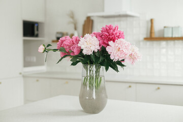 Beautiful peonies in vase on granite countertop on background of stylish white kitchen with wooden shelves and appliances in new house. Modern minimal kitchen interior