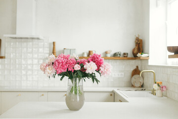Beautiful peonies in vase on granite countertop on background of stylish white kitchen with wooden shelves and appliances in new house. Modern minimal kitchen interior - obrazy, fototapety, plakaty