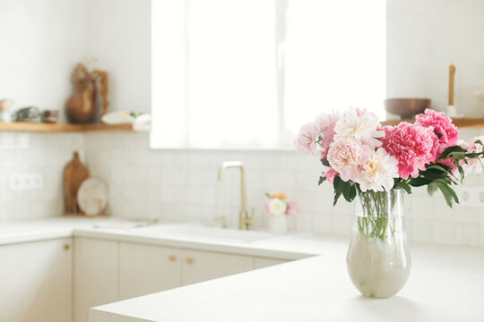 Beautiful peonies in vase on granite countertop on background of stylish white kitchen with wooden shelves and appliances in new house. Modern minimal kitchen interior