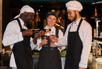 Two happy young intercultural cooks in uniform holding wineglasses while African American male colleague pouring sparkling champagne