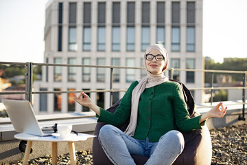 Carefree young female wearing hijab and glasses meditating in bean bag chair with hands in gyan...