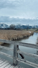 The Hopfensee in winter photographs from a hiking trail on a wooden bridge with dramatic sky and snow covered mountains