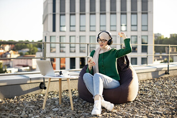 Emotional muslim lady in hijab singing along musical phrases in headphones while resting in pouf chair on terrace. Excited young woman rocking out to energetic tunes of favourite melodies outdoors.