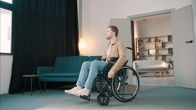 Young caucasian man sitting in wheel chair in his room looking seriously out the window