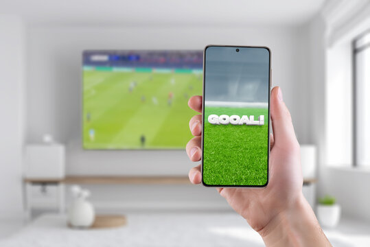 Goal on the phone display. The concept of tracking sports results online. Living room with a TV and soccer game in background