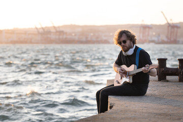 A man with curly hair wearing wireless headphones sits at the quay and playing guitar at early sunset