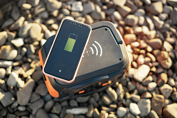 Portable digital device with green battery status on screen placed on multitasking power station outside buildings. Cordless charging of mobile phone fixing problems of location and cable chaos.