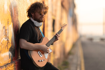 Romantic man hipster wearing sunglasses standing by the wall and playing guitar at early sunset