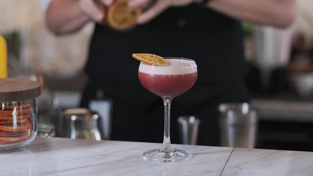 A bartender in an expensive edith hotel pours a prepared pink cocktail from a shaker into a glass and decorates with a slice of lemon