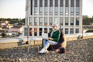 Wide view of arabian woman in traditional headscarf working on laptop while sitting in soft chair in fresh air. Discharged gadget being connected to renewable energy source on panoramic terrace.
