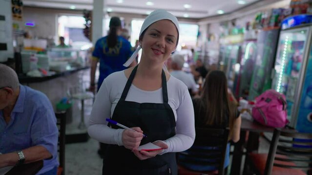 Portrait of waitress standing inside restaurant cafeteria looking at camera smiling. Female server holding pen and notepad ready to take customer orders