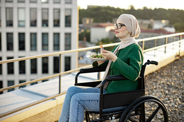 Beautiful arabian woman wearing glasses and hijab holding plate full of vegetables while enjoying cityscape in wheelchair. Confident young female in jeans tasting healthy food in open air on rooftop.