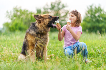 Cute little girl eating ice cream and feeding it to her shepherd dog sitting on the grass in the park in summer. High quality photo, blurred background.