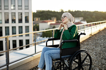 Pretty bespectacled woman with mobility issues daydreaming with hands under chin in sunlight on roof terrace. Relaxed muslim person wearing jeans and hijab soaking up with vitamin D outdoors.