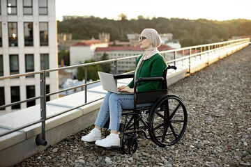 Focused young female with mobility impairment using portable computer while staying in open air on roof. Professional arabian entrepreneur in hijab analyzing new project via modern technologies.