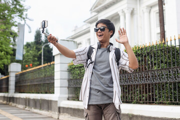 Happy excited young travelerman recording video travel vlog on urban sidewalk greeting with hand.