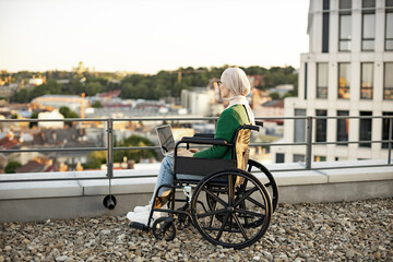 Back view of arabian person in traditional headscarf staring into distance on rooftop against urban background. Relaxed female wheelchair user with laptop admiring picturesque scenery outside.