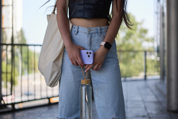 Fashionable young girl with a shopper bag on shoulder, modern smart and glass bottle of water in hands. Trendy female person posing in the city street