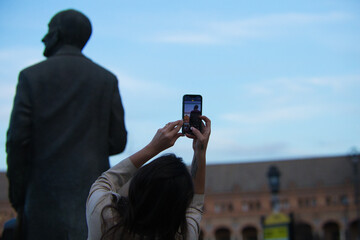 Woman taking a photo with her mobile phone of a statue in the most famous square of seville in spain. in the mobile phone you can see the monument and the statue from behind.