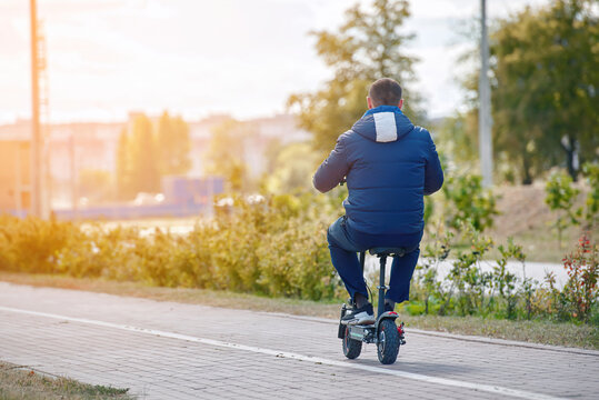 Man Sitting On Electric Scooter And Driving Through City Road. Alone Man In Warm Clothes Ride Electro Scooter In The City In Autumn Season. Electric Bike - Personal Mobility Vehicle