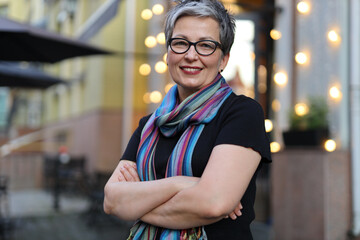 Fototapeta premium An adult woman with a short hairstyle and glasses in front of a street cafe with blurry lights.